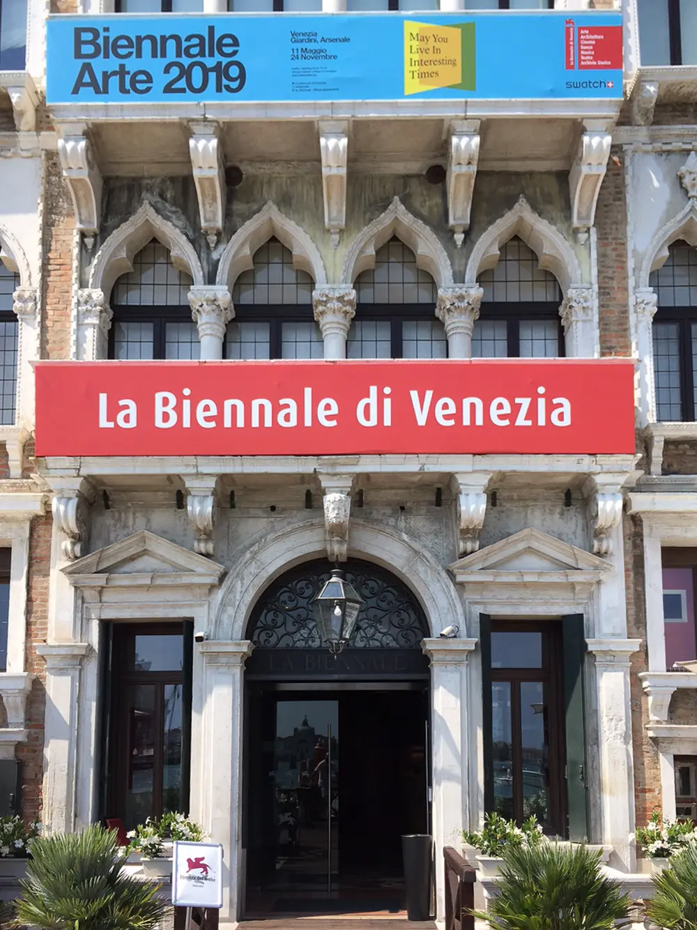 The arched Gothic entrance of an old Venetian palace, with a red banner reading La Biennale di Venezia and a blue and yellow sign for Biennale Arte 2019 above it.