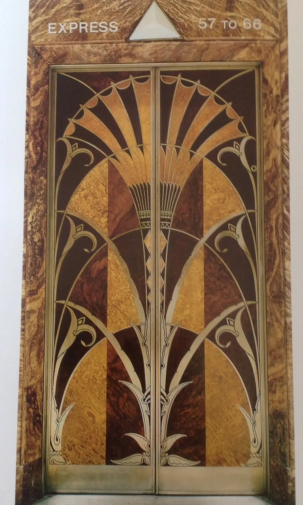 A pair of tall elevator doors covered in inlaid woods of several colours, forming a symmetrical fan of thin, pointed, plant-like shapes that rise from the bottom, inside a metal frame with a triangle above.