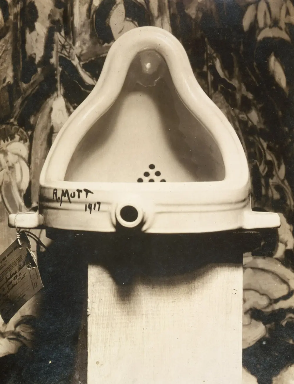 A black-and-white photograph of a white porcelain urinal turned onto its back on a plinth, signed "R. Mutt 1917" in black paint, in front of a dark painting.