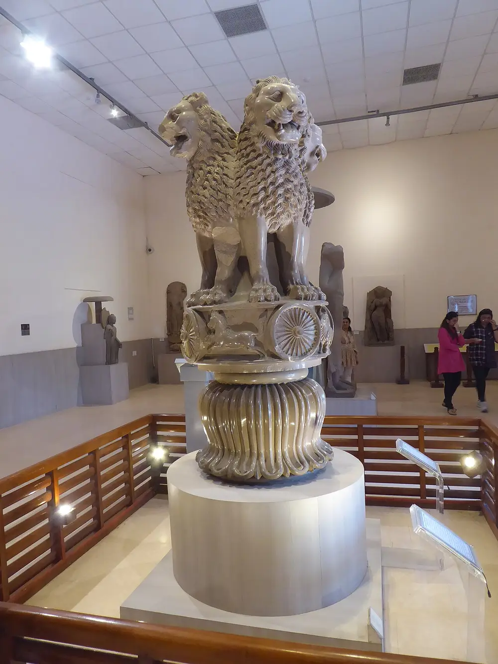The Lion Capital in a museum hall: four stone lions sitting back to back on a round base carved with a horse and a spoked wheel, above a bell-shaped lotus, on a round pedestal behind a wooden railing.