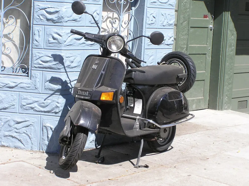 A dark grey Bajaj Chetak scooter with a rounded front shield, a long black seat and a spare wheel at the back, parked on a pavement in front of a pale blue patterned wall.