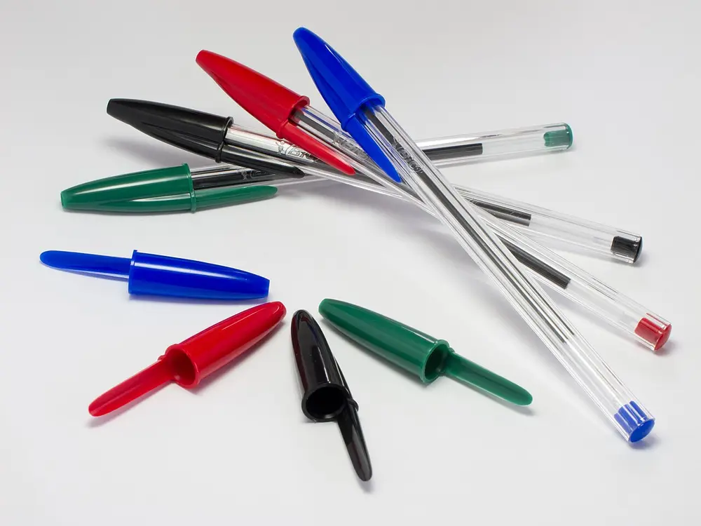 Four clear, six-sided Bic Cristal ballpoint pens lying on a white surface with their blue, red, black and green caps beside them.