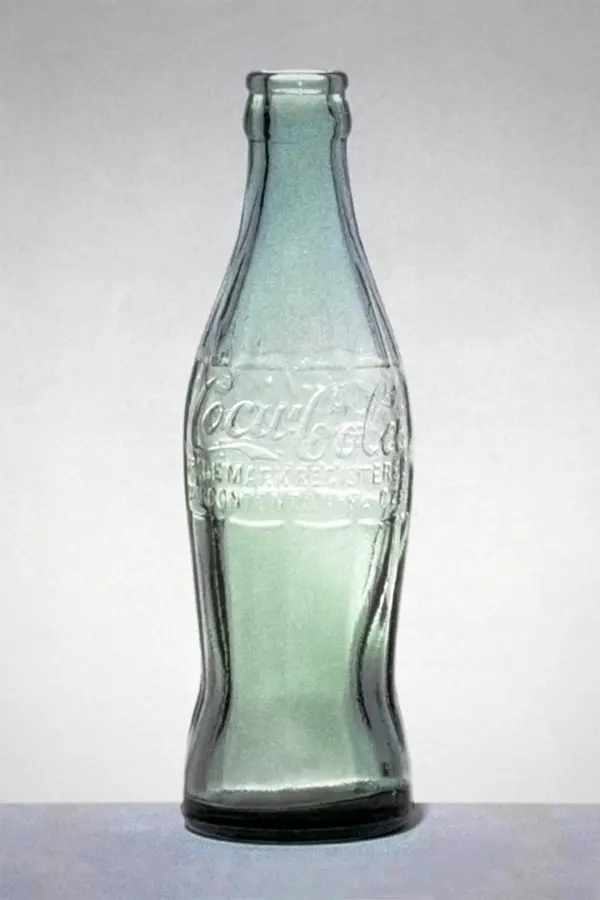 A pale green glass Coca-Cola bottle with fine vertical ribs and the Coca-Cola name moulded into the glass, narrow at the neck, bulging in the middle and pinched near the base.