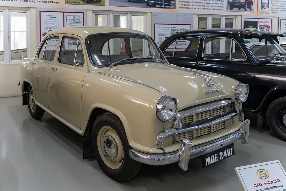 A cream Hindustan Ambassador Mark I car with a rounded body, a chrome grille and bumpers and round headlamps, on display in a car museum.