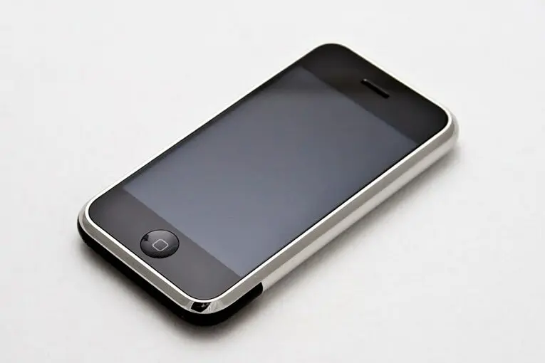 The first iPhone lying at an angle on a white surface, with a black glass screen filling the front, one round button below it and a silver metal edge.