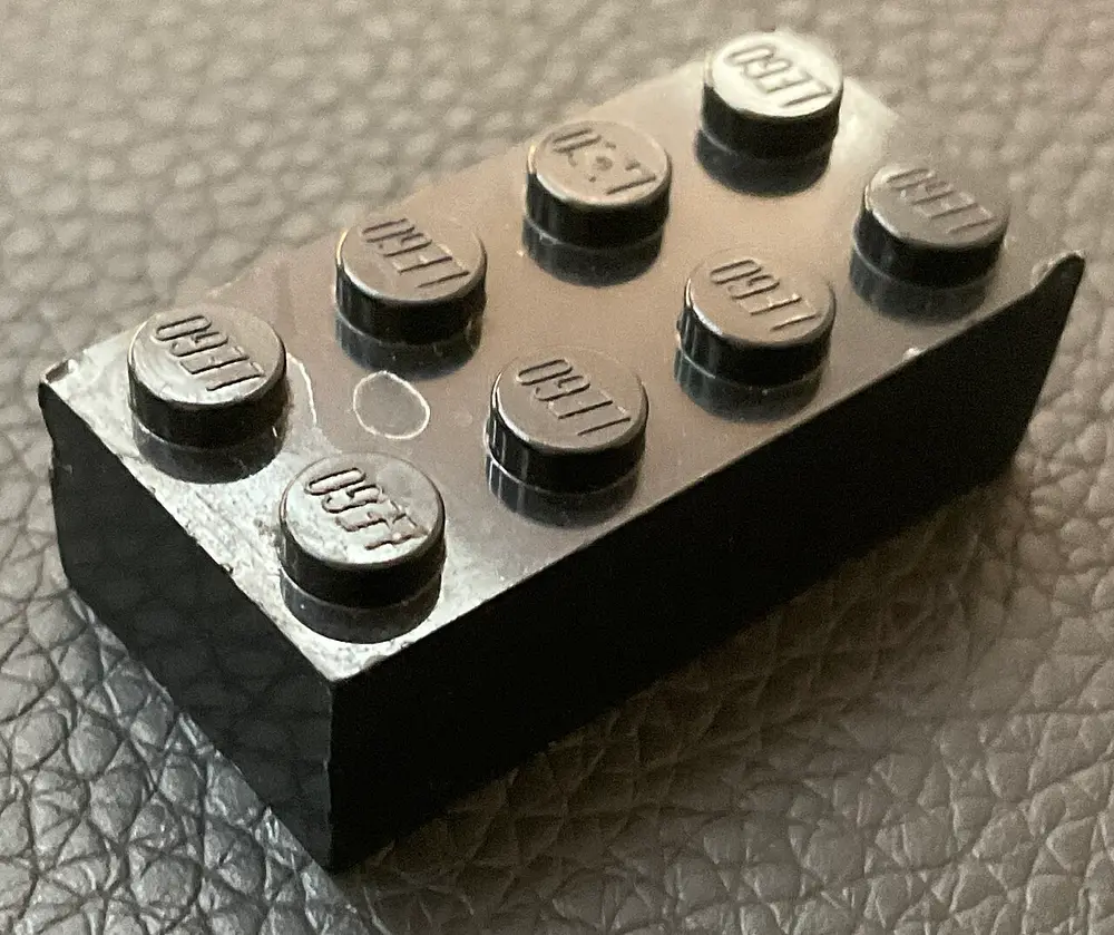 A single black Lego brick with two rows of four round studs on top, each stud stamped with the word LEGO, resting on a grey leather surface.