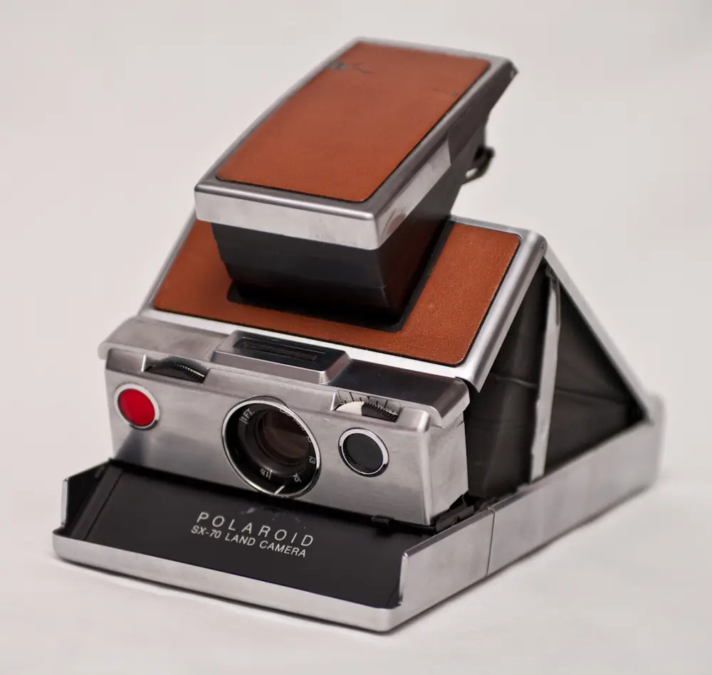 A Polaroid SX-70 camera opened up into its triangular shape, with a brushed silver metal body, brown leather-look panels, and the lens and a red shutter button on the front.