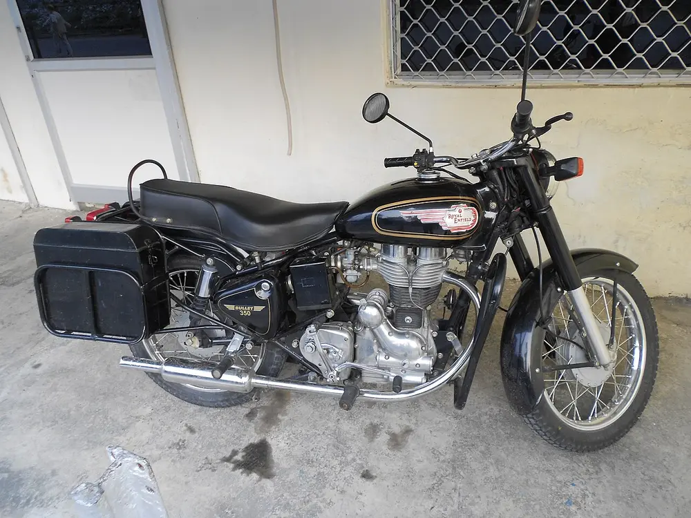 A black Royal Enfield Bullet 350 motorcycle with a chrome engine, a round headlamp and black saddlebags, parked in front of a white building with a barred window.