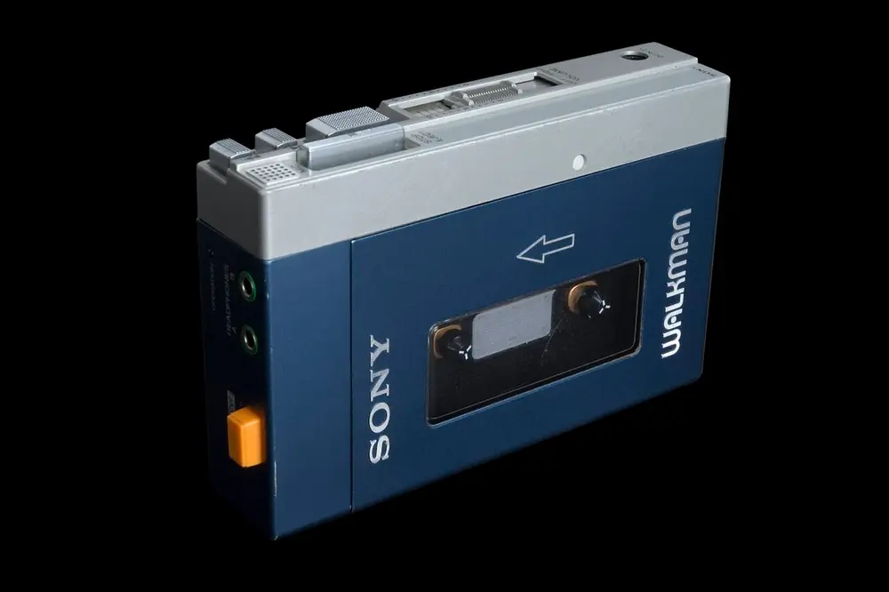 A blue and silver Sony Walkman cassette player with a window showing the tape inside, an orange button on the side and the word WALKMAN on its edge, photographed against a black background.