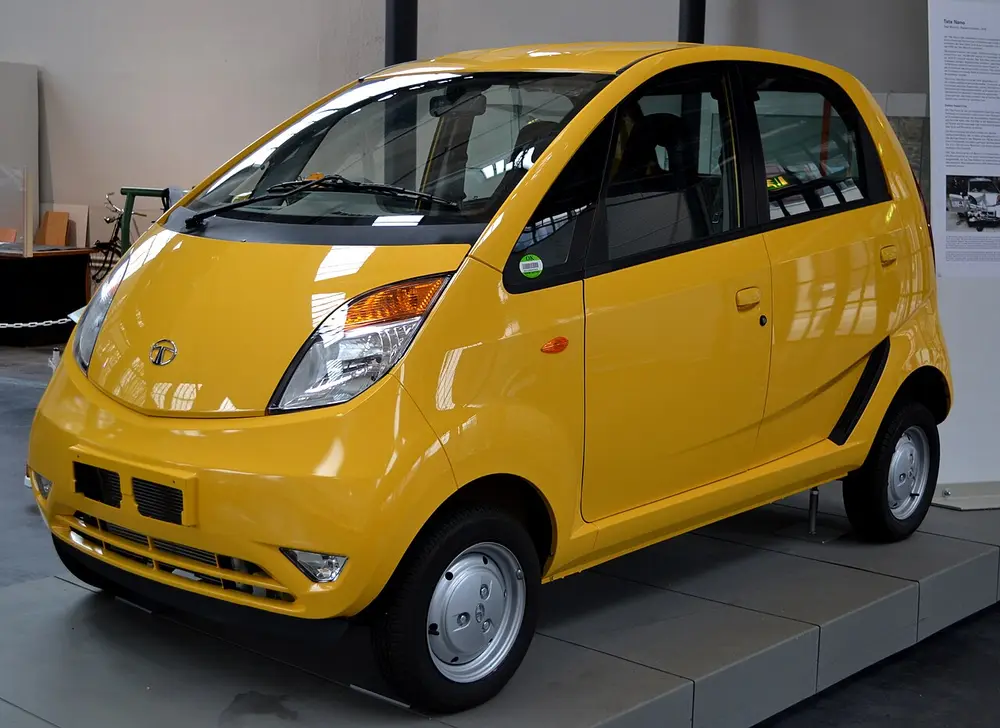 A small, tall, rounded bright yellow Tata Nano car with big headlamps, on display in a museum hall.