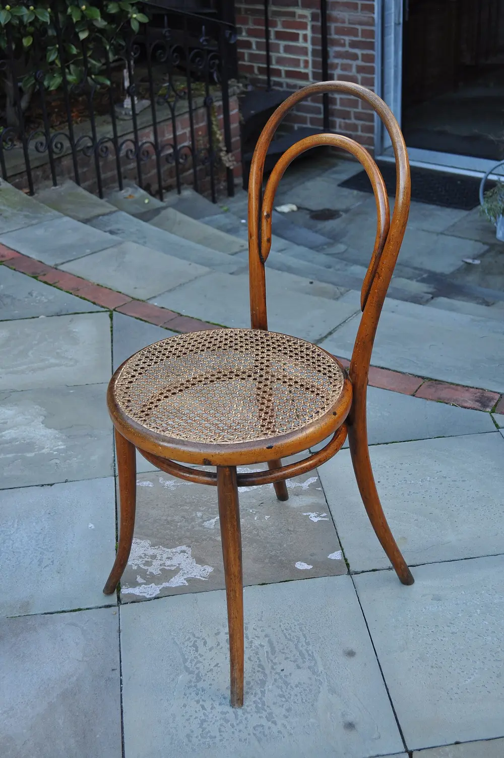 A dark brown bentwood chair with a round woven cane seat, a back made of two curved wooden loops, and a wooden ring joining the four legs, standing on a stone pavement.
