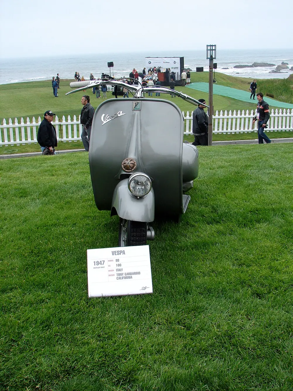 A grey 1947 Vespa scooter seen from the front on a lawn at an outdoor show, with its rounded front shield and single headlamp, and a small information sign in front of it.