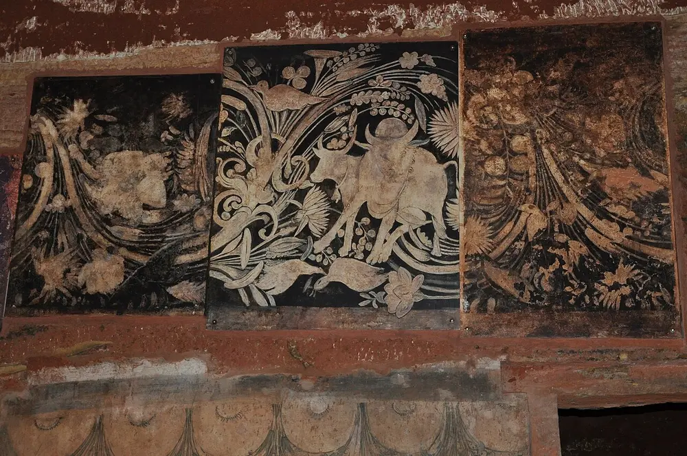 Three dark painted panels set on a red cave wall, the middle one showing a pale bull among curling leaves, flowers and birds.