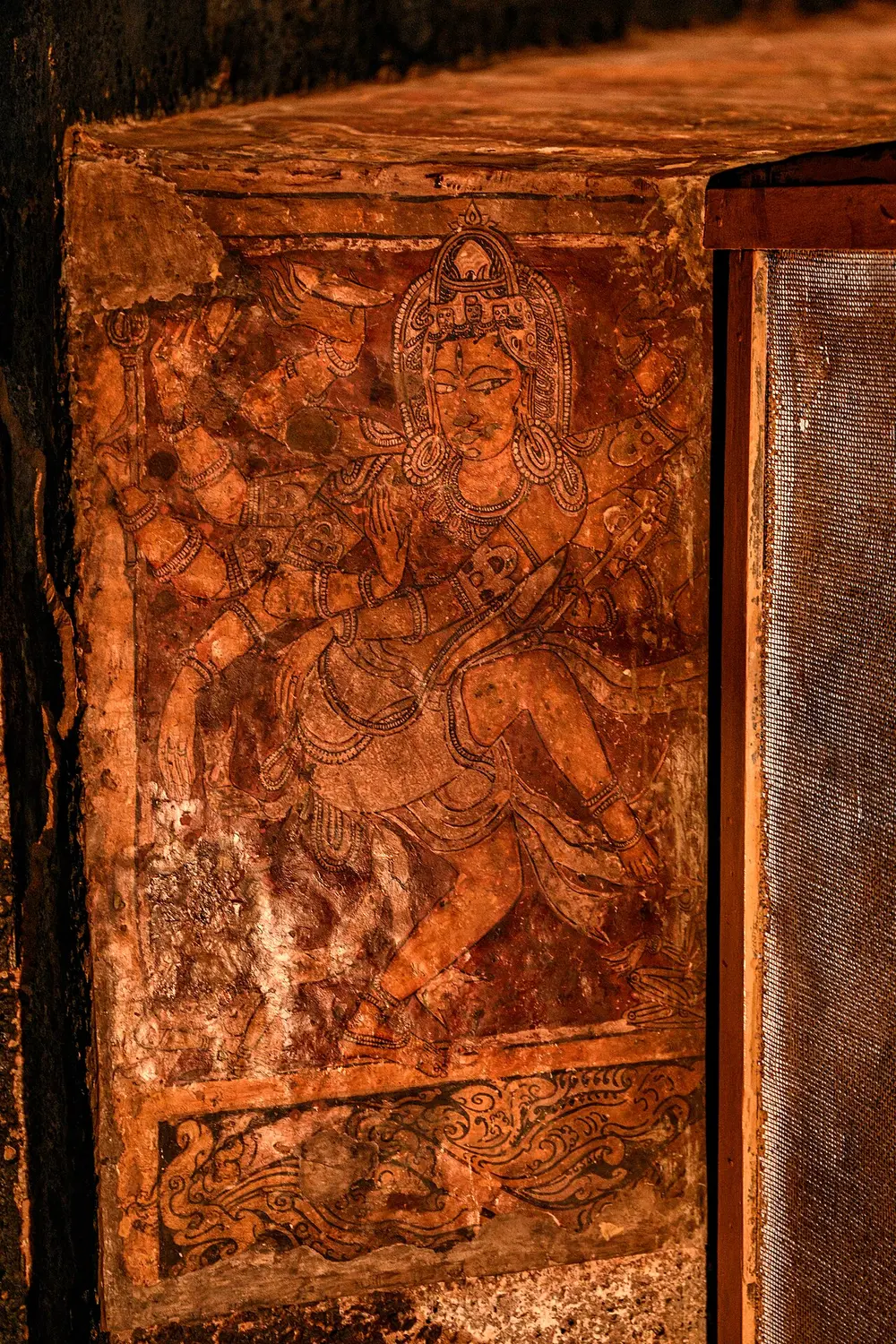 A faded red-brown wall painting of a many-armed god dancing with one leg raised, wearing a tall crown, drawn with fine dark outlines, above a band of curling waves.