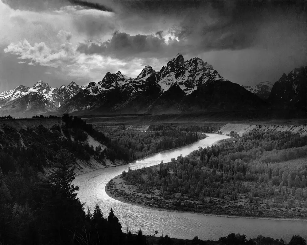 A black and white landscape of a river curving in great S-bends through a dark forested valley towards a range of jagged, snow-streaked mountains under a bright, cloudy sky.