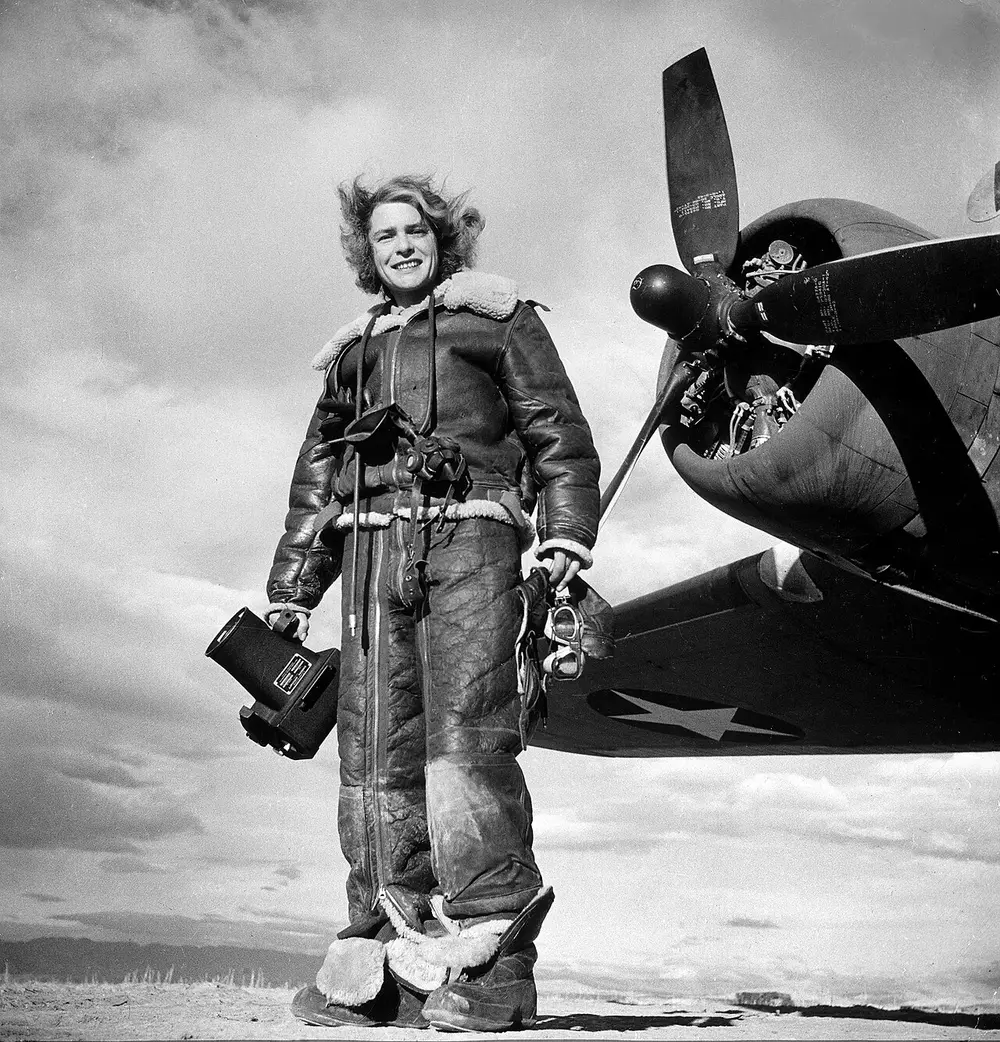 A black and white photo of a woman in a fur-lined flying suit and boots, holding a large camera, standing in front of the propeller of a military plane.