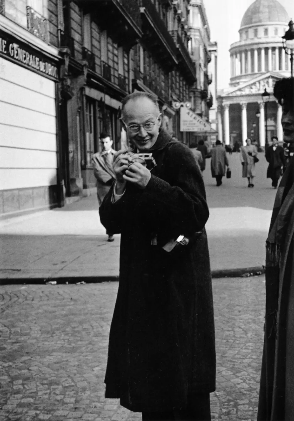 A black and white photo of a man in a long dark coat on a Paris street, holding a small camera up to his face, with the dome of the Panthéon behind him.