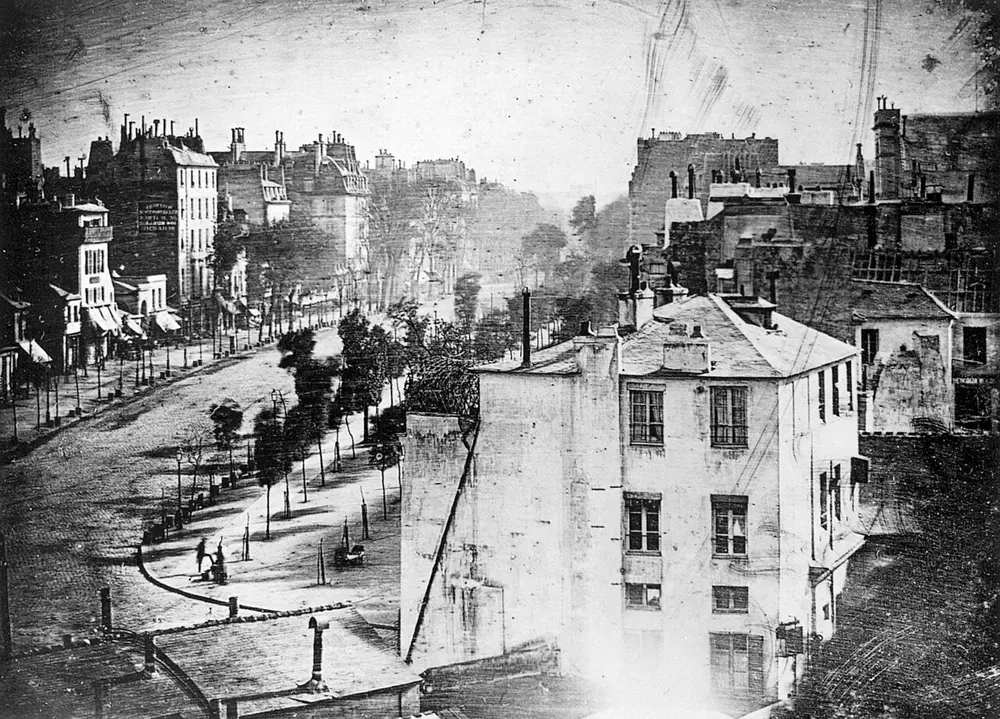 A sharp black and white view down a long, tree-lined Paris street from a high window, with tall buildings and rooftops but almost no traffic. A tiny standing figure is just visible on the pavement at the lower left.