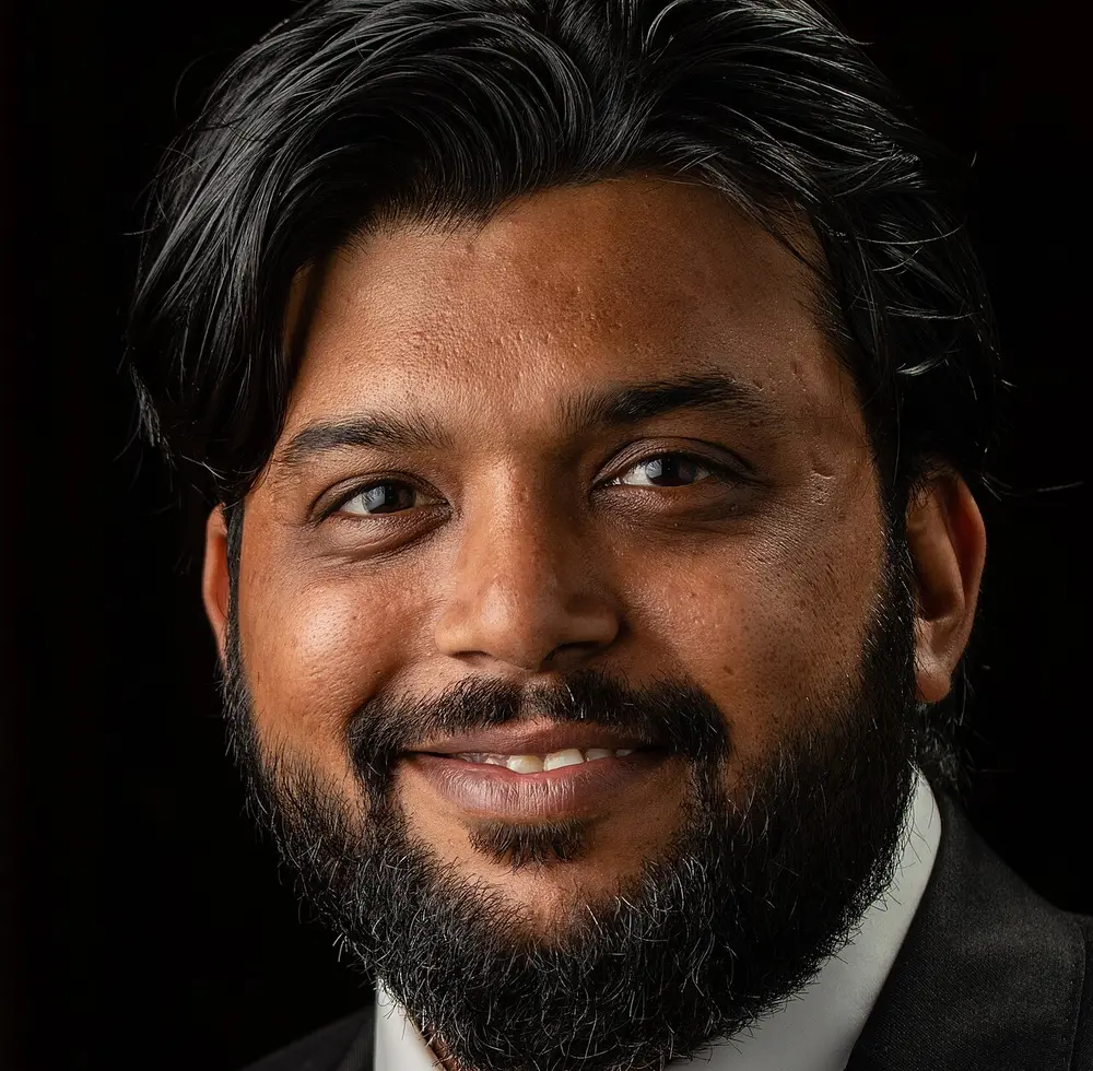 A smiling man with dark wavy hair and a beard, in a dark suit and white shirt, against a black background.