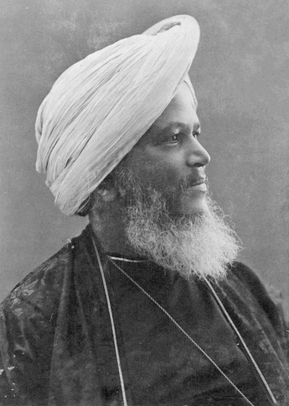 A black and white portrait of an elderly man with a white beard and a tall white turban, wearing a dark coat, looking to one side.