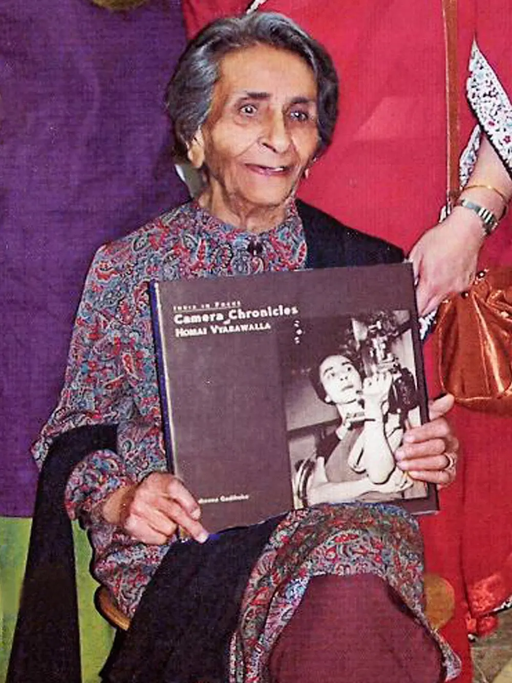 An elderly woman in a patterned sari sits holding a large book of her black and white photographs, against a red and purple background.