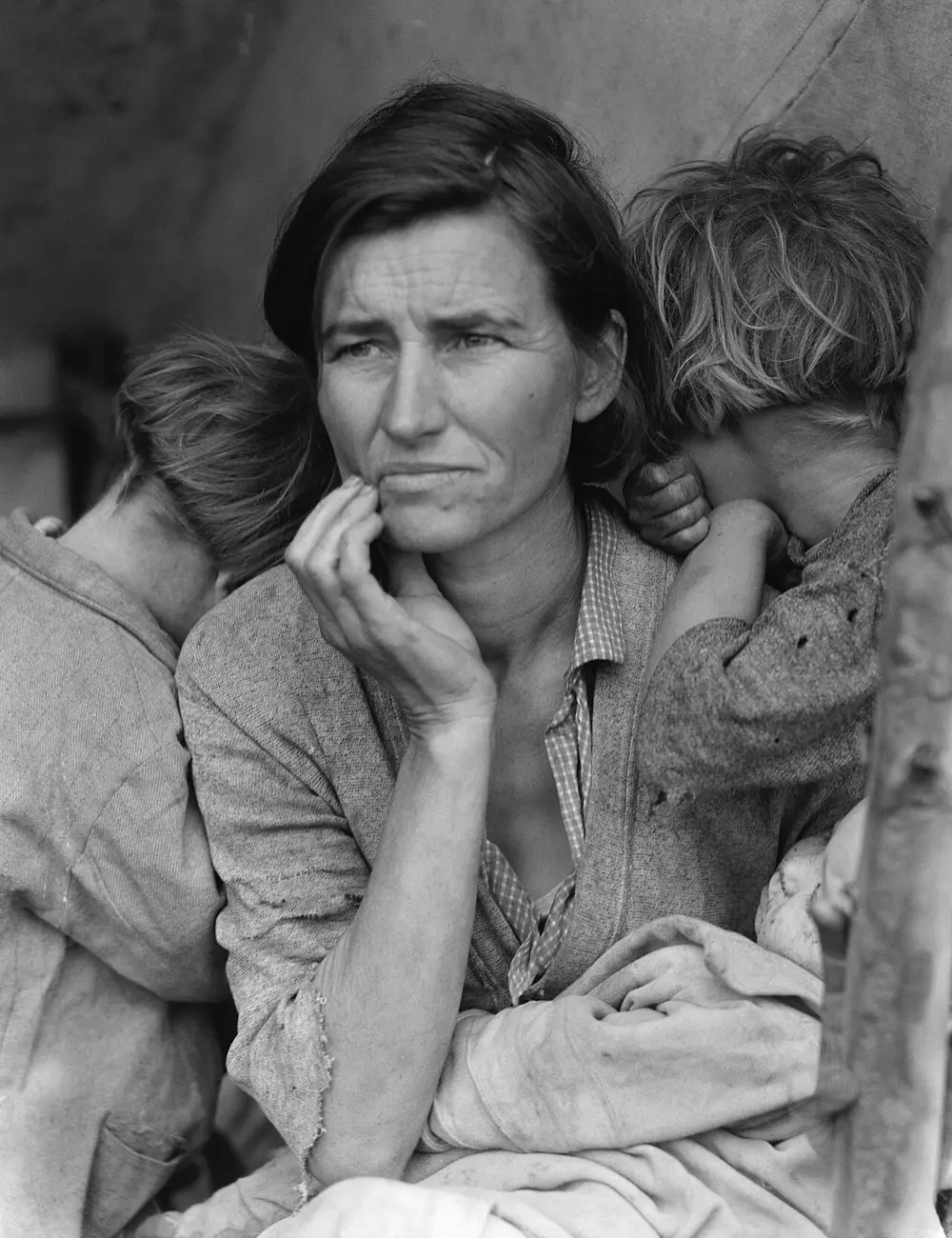 A black and white photo of a worried woman with her hand at her chin, gazing into the distance, with two children leaning on her shoulders with their faces turned away and a baby on her lap.