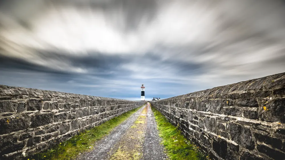 A gravel path runs straight between two low stone walls towards a small black and white lighthouse in the distance, under a sky of streaked grey clouds.