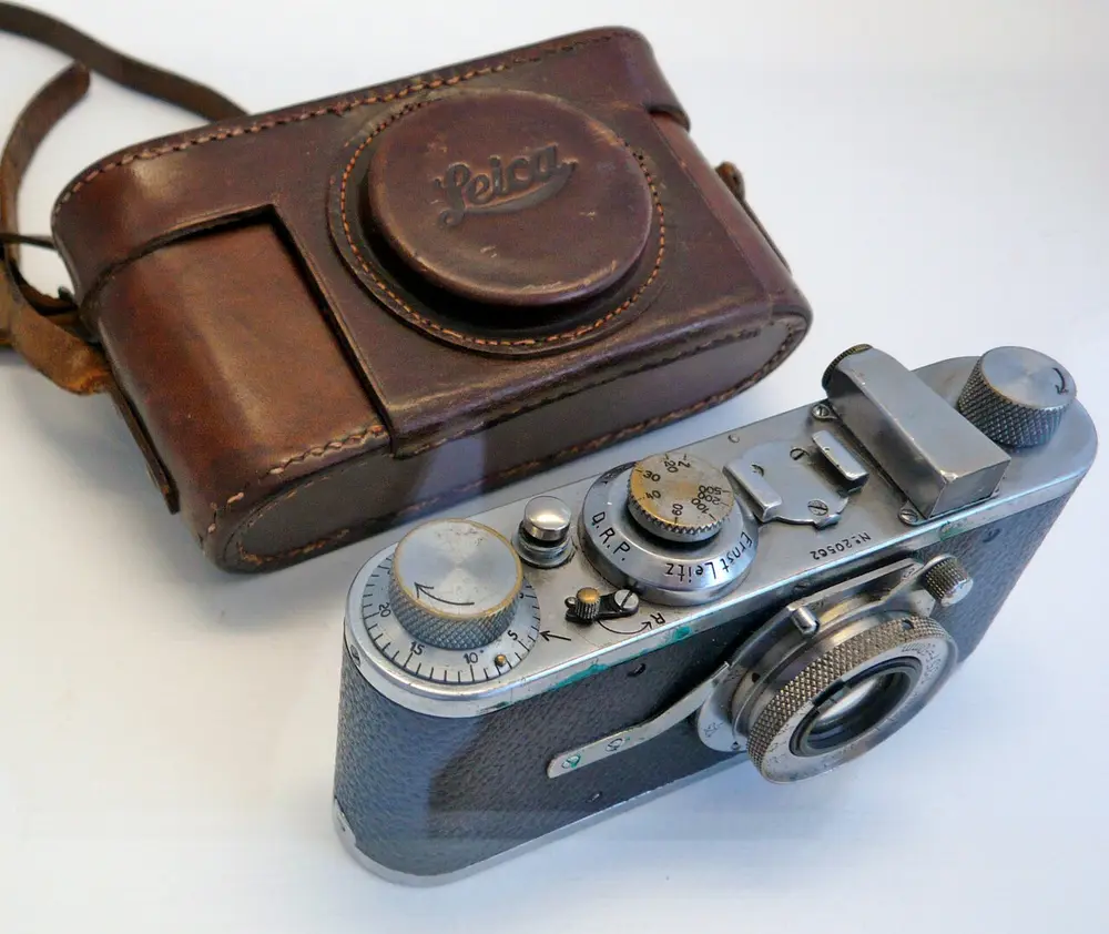 A small silver and black camera with round dials on top and a short lens, lying in front of its worn brown leather case.