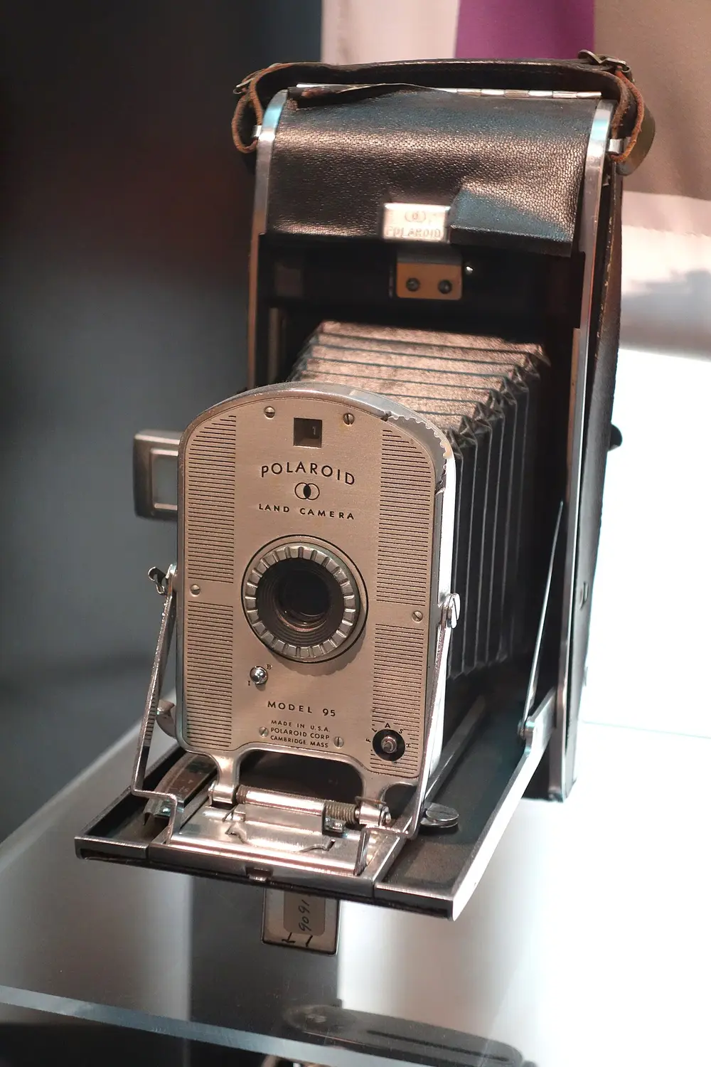 A folding camera with a leather-covered body, black bellows and a round lens on a fold-out front, standing open in a museum case.
