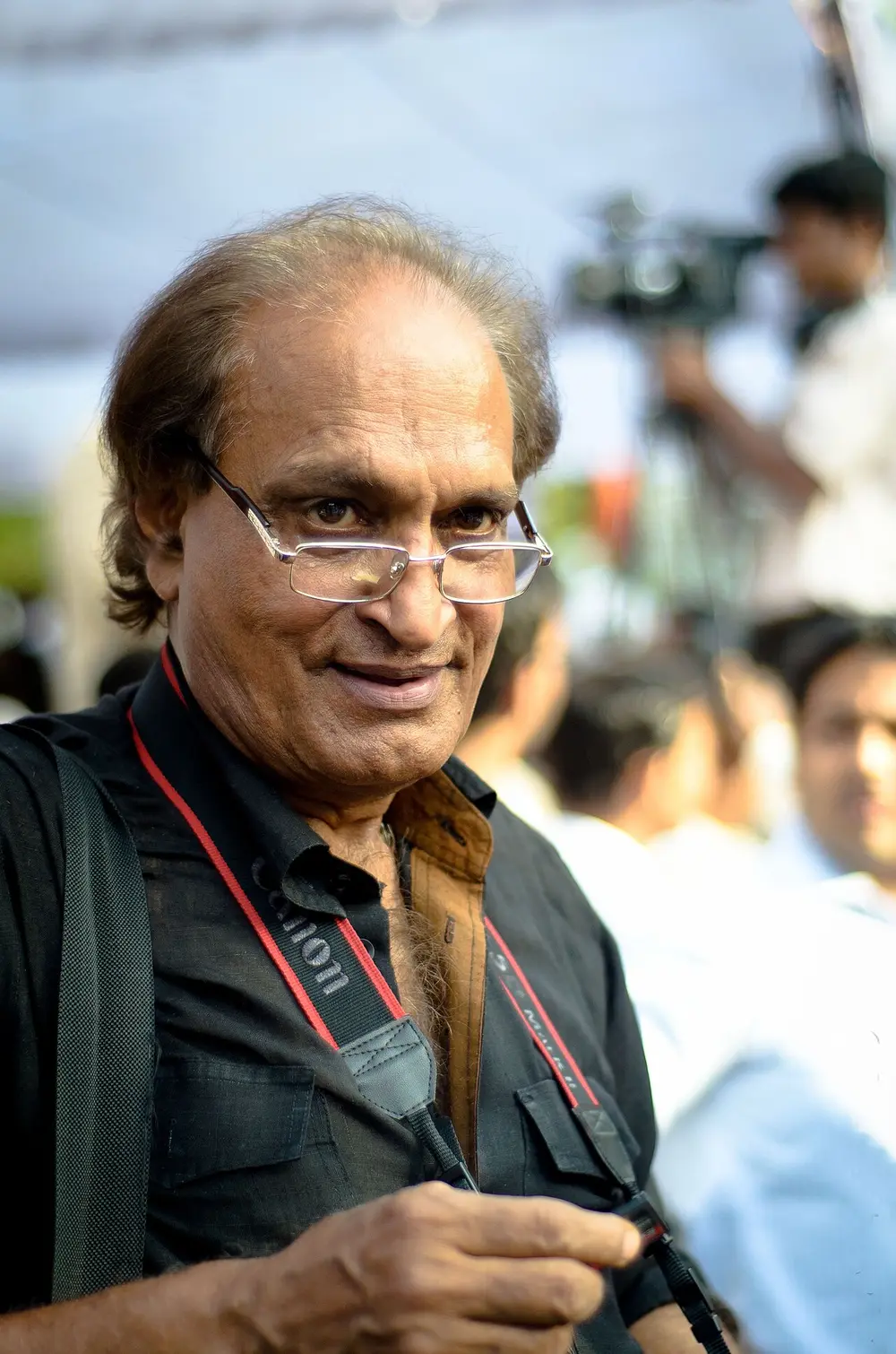 A smiling man with glasses and grey hair, a camera strap round his neck, at work among a crowd outdoors, with a television camera behind him.