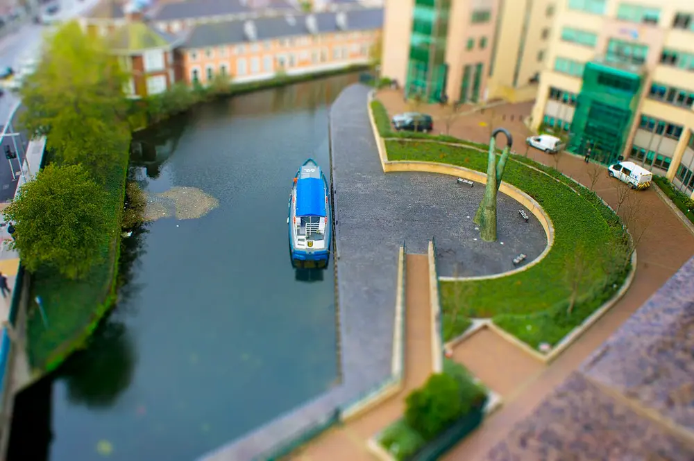A canal seen from high above, with a blue and white boat, a curved paved path and trees. Only a band across the middle is sharp and the top and bottom are blurred, so it looks like a model railway scene.