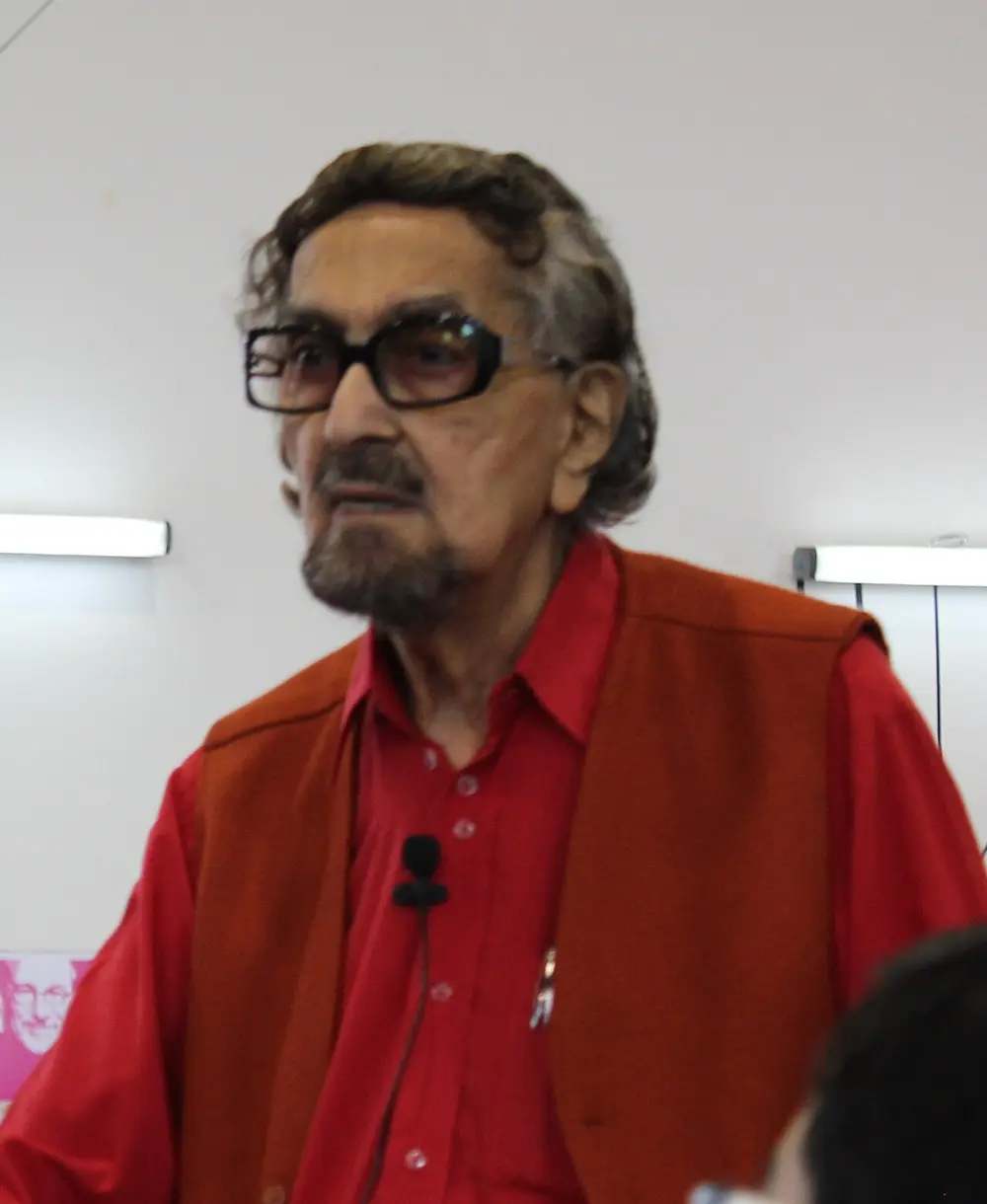 A photograph of Alyque Padamsee as an older man with grey hair, a grey beard and dark-framed glasses, in a red shirt and red waistcoat, with a small microphone clipped to his shirt.