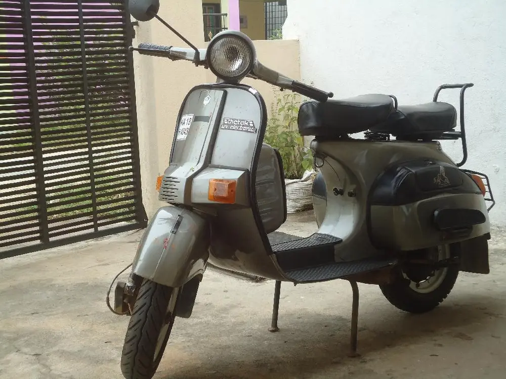 A restored grey Bajaj Chetak scooter parked on its stand beside a wall, with a round headlamp, a curved front shield and a long black seat.
