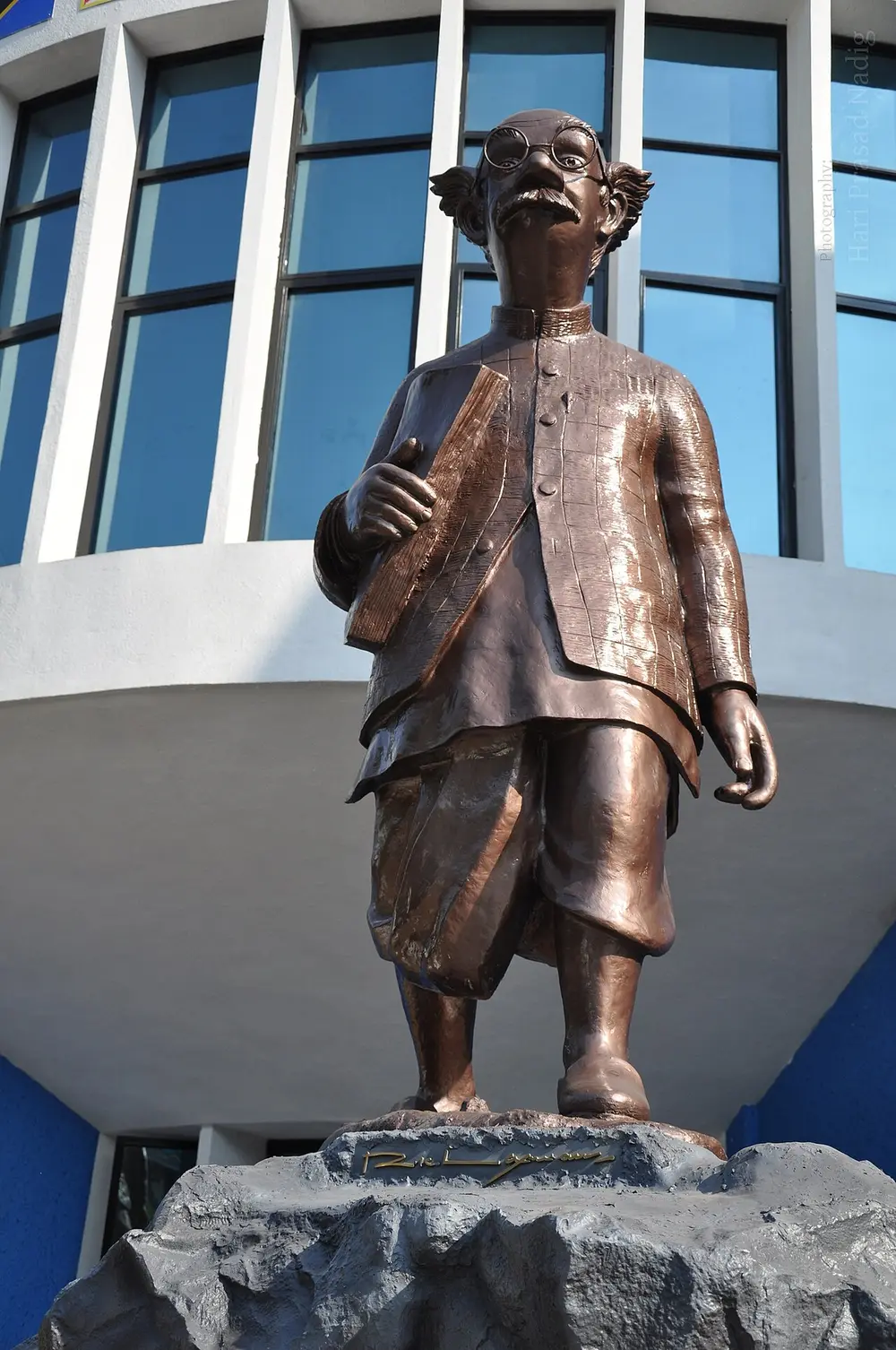 A tall bronze statue of a small, bald old man with round glasses, tufts of hair above his ears, a checked jacket and a dhoti, standing on a rough stone base in front of a building with blue glass windows.