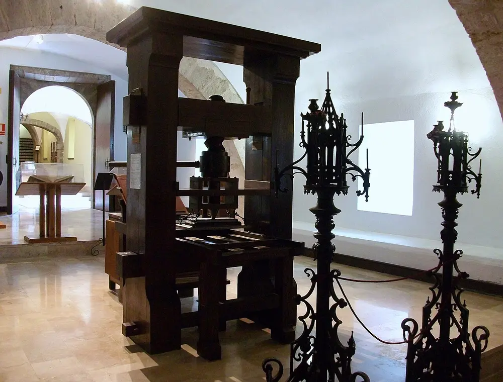 A tall, dark wooden hand press with a heavy upright frame and a big central screw, standing in a museum room with white arched walls beside two ornate black iron stands.