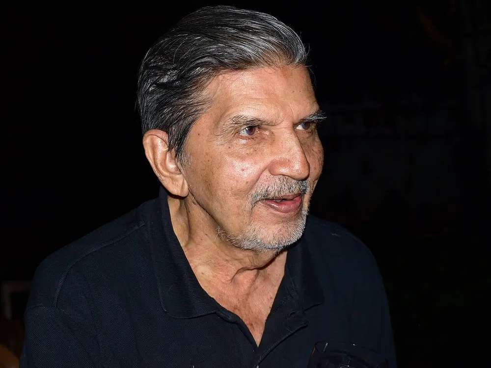 A photograph of Mario Miranda as an older man with swept-back grey hair and a short grey beard, smiling, in a dark shirt against a black background.
