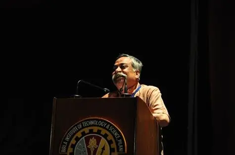 A photograph of Piyush Pandey, small in the frame, speaking into a microphone at a wooden podium on a dark stage.
