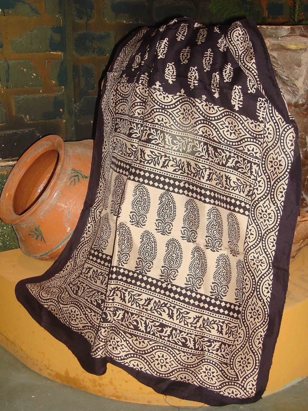 A silk stole hanging over an orange clay pot, printed in black on a cream ground with rows of paisleys and bands of small flowers and dots.
