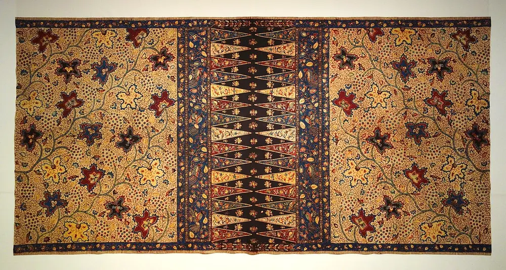 A long brown and cream cloth in a museum, covered with dark red and blue flowers on vines, with a band of tall triangles down the middle.