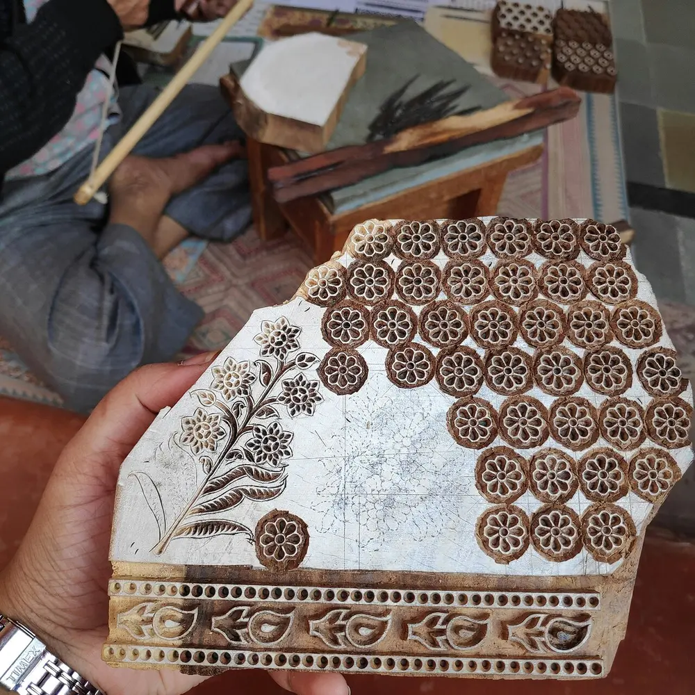 A hand holding a half-finished wooden printing block, its face carved with rows of small flowers and a leafy sprig, with a carver's tools behind.