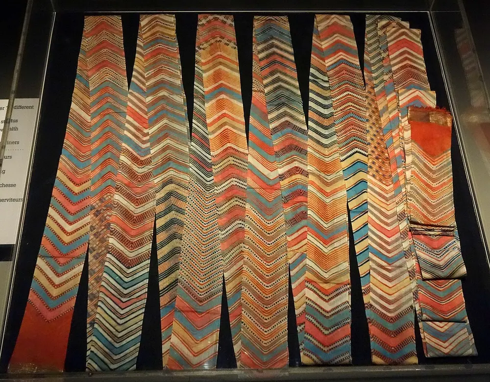 Several long cloths laid side by side in a museum case, dyed in zigzag bands of orange, blue, cream and green.