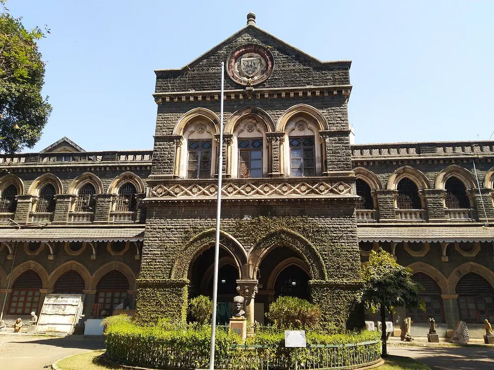 A grey stone building in Gothic style with pointed arched windows, a gabled front and a round clock-like emblem, behind a garden.