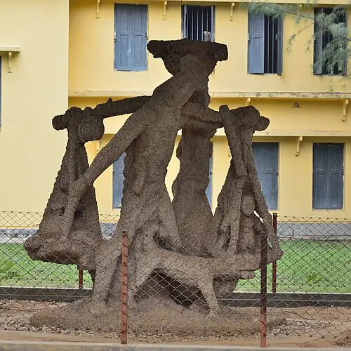 A large rough grey outdoor sculpture of a man carrying a pole on his shoulder with a basket hanging at each end, a woman walking beside him and a dog at their feet, in front of a yellow building.