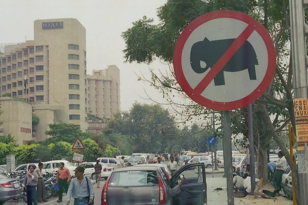 A round road sign with a red border and a red slash across a black elephant, on a busy New Delhi street, with a triangular pedestrian sign and a blue round sign in the background.