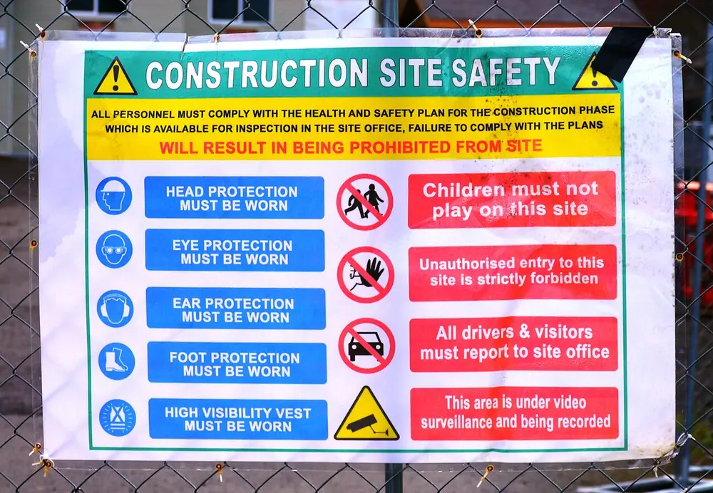 A building site notice with blue circles showing a hard hat, goggles, ear defenders, boots and a safety vest on the left, and red circles with slashes over children, a hand and a car on the right, plus a yellow triangle with a camera.