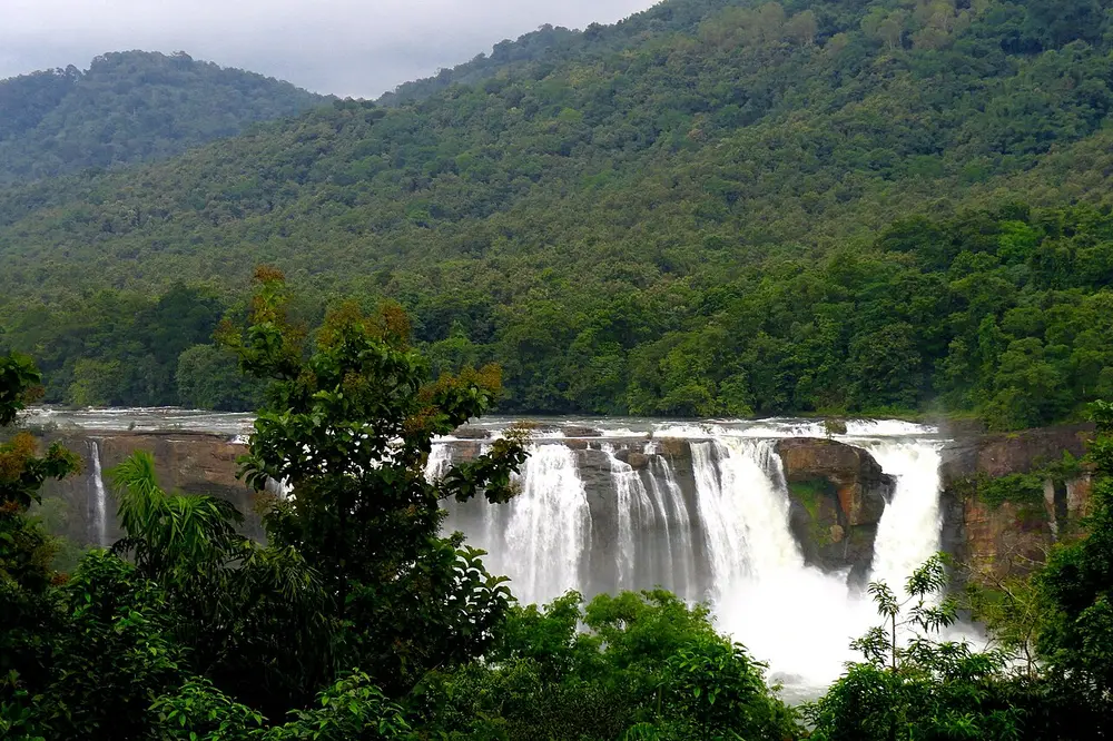 A broad white waterfall pouring over a rock ledge in the middle of thick green forested hills.