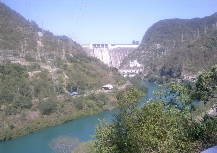 A tall concrete dam wall filling a narrow gap between rocky hills, with power lines and a river below.