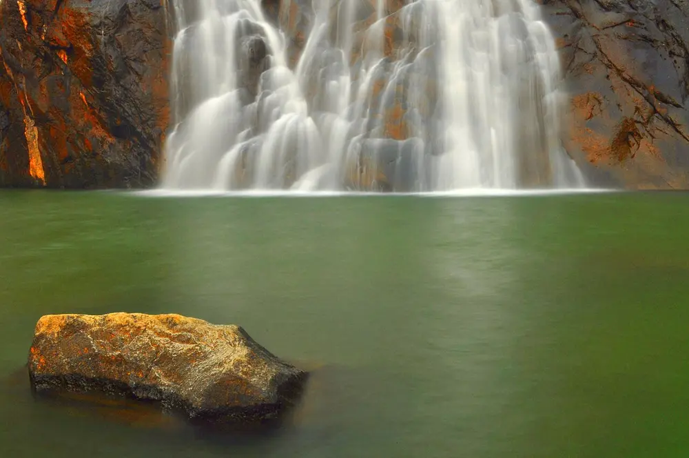 A white curtain of water falling down an orange rock face into a calm green pool.