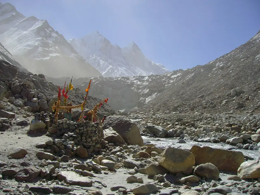 A grey wall of glacier ice at the end of a stony valley, with snowy peaks behind and prayer flags on a cairn of rocks in front.
