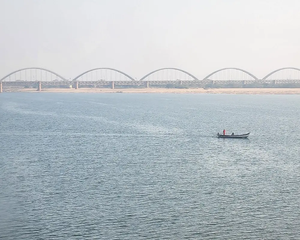 A very wide river with a small fishing boat, and a long bridge of repeated arches on the horizon.
